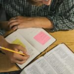 A Man Writing on a Notebook while Reading a Bible